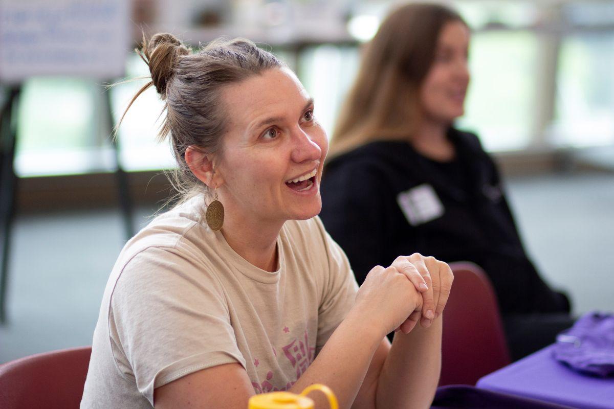 Women sits at table with someone in the background and smiles at someone coming up to the table