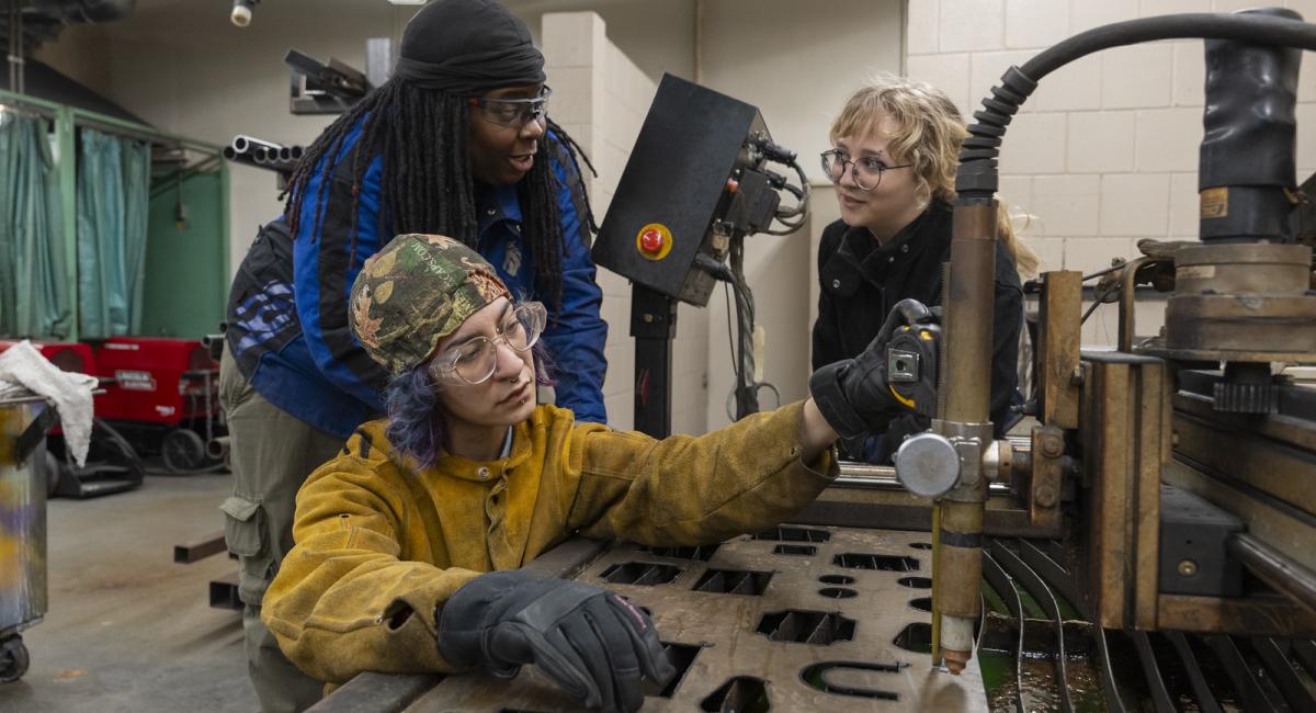 Woman welding with two other woman in the back working on welding projects