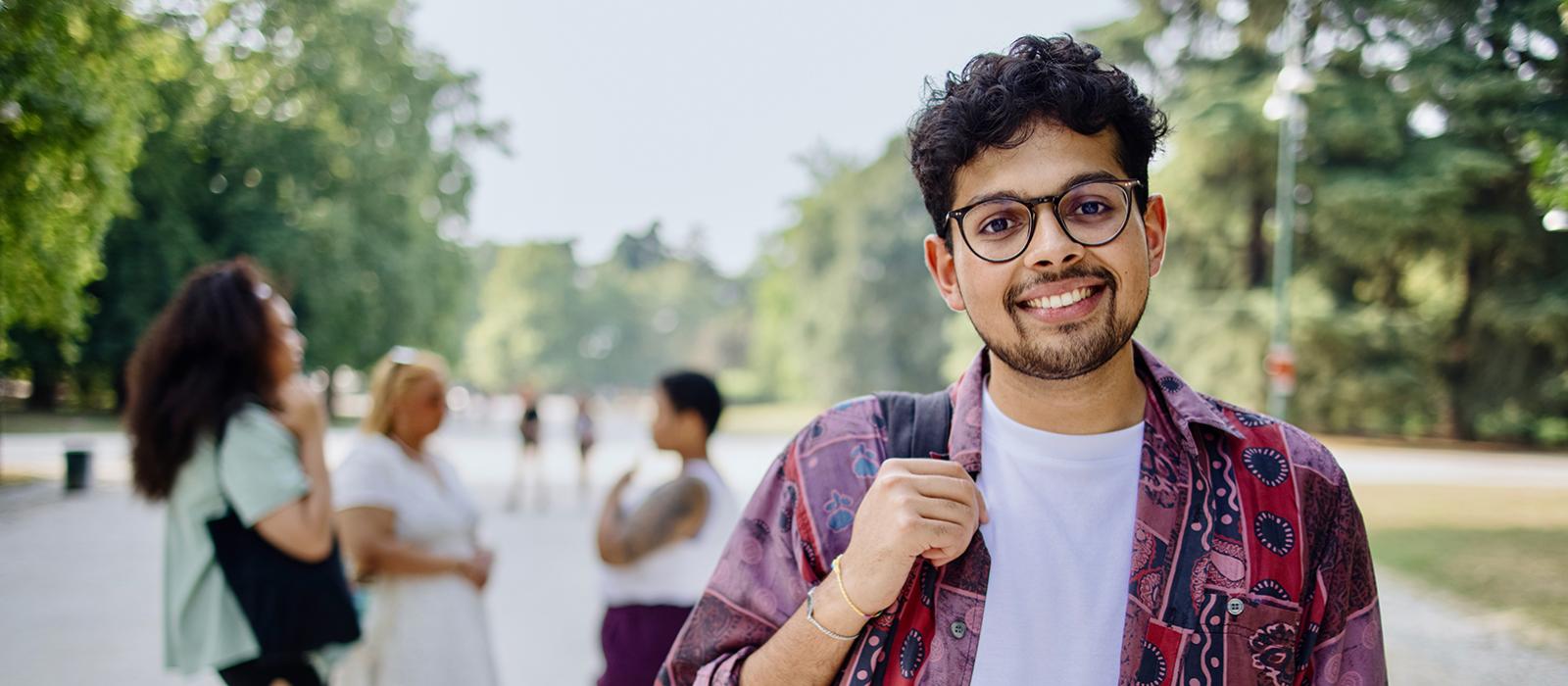 A male smiling male student with a backpack stands outside on campus.