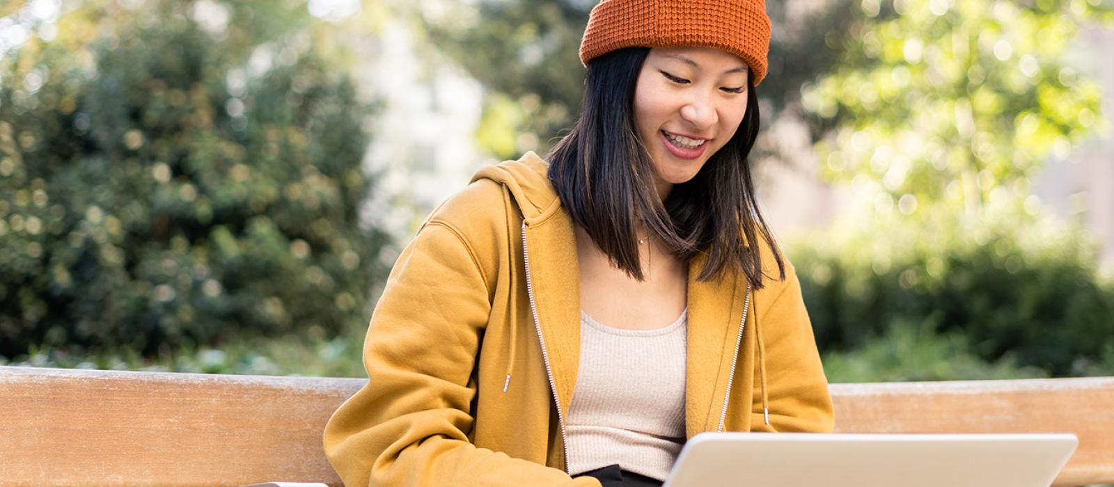 Smiling student in a winter hat working on a laptop outside in the fall