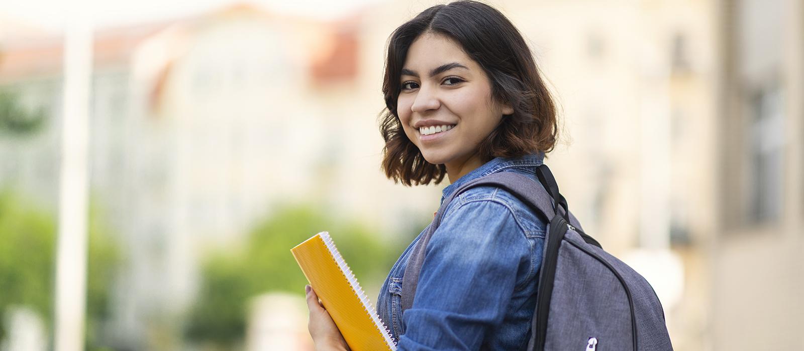 A student in a jeans jacket with a backpack looks over her shoulder, smiling. 