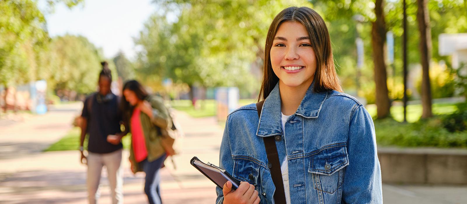 A female student outside in the park carrying a notebook smiling. 