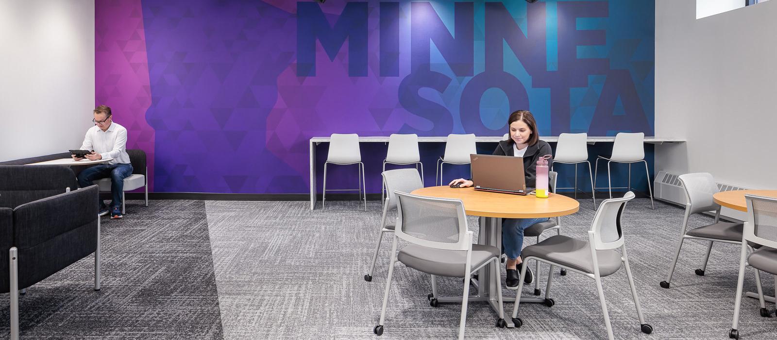 Two students study at separate tables in the Management Education Center in front of a branded wall that says, "Minnesota."