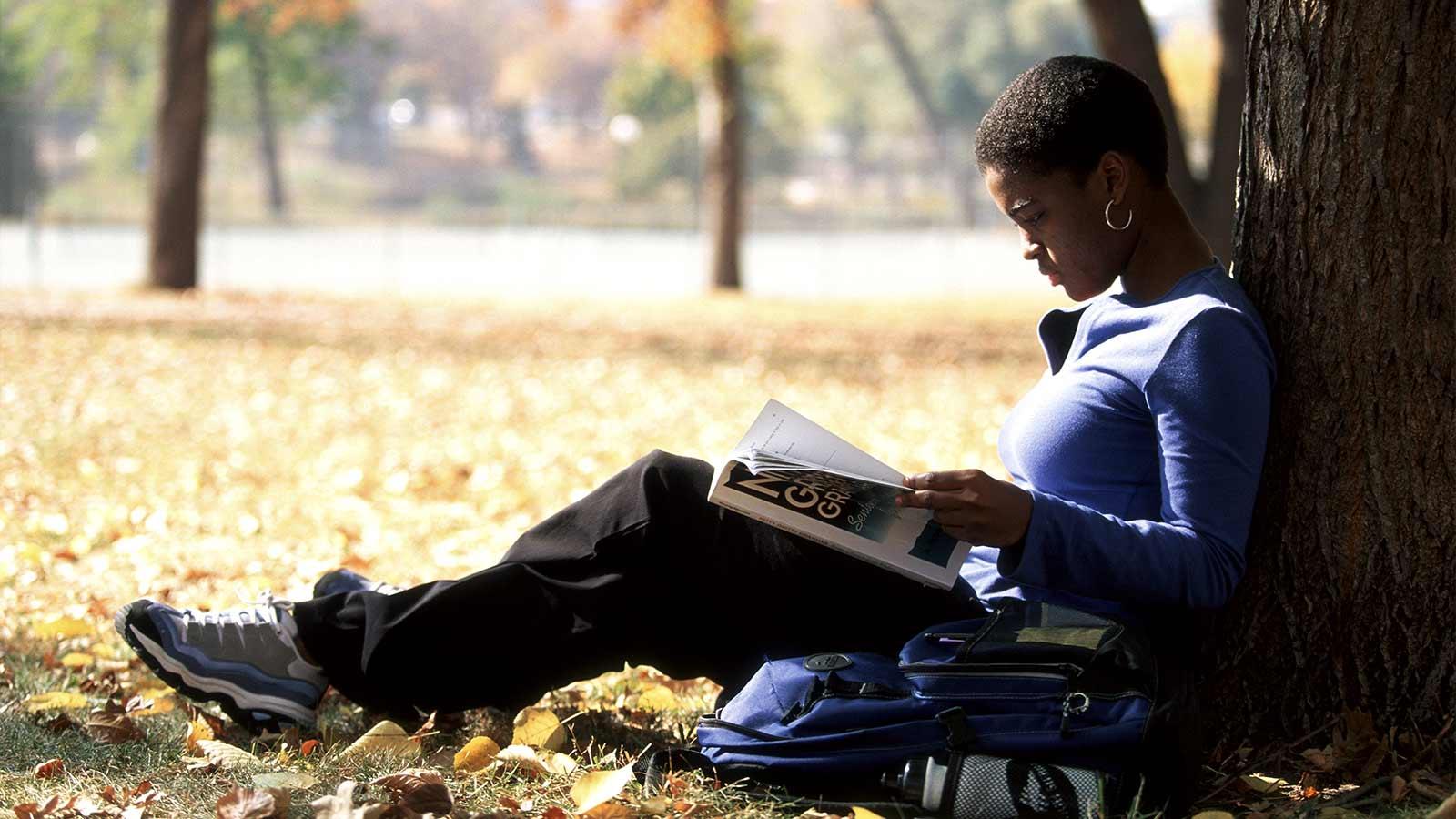 student reading a textbook while leaning against a tree.