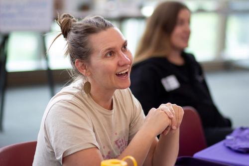 Women sits at table with someone in the background and smiles at someone coming up to the table