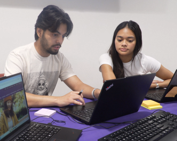 MAle student sits at a table with a laptop in front of him and smiles for the camera