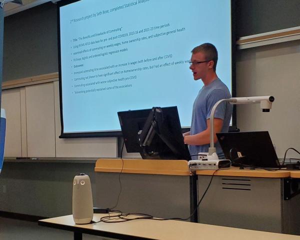 Student presents in front of  classroom with a large projector screen behind him. 