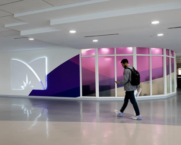 A student, looking at their phone walks past a large illuminated M logo mark. 