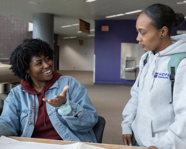 Two women together with one sitting at a table explaining something to the other who is standing up and smiling