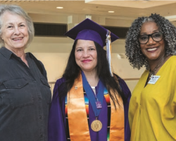 Commencement speaker stands in cap and gown next to two women and smile for the camera