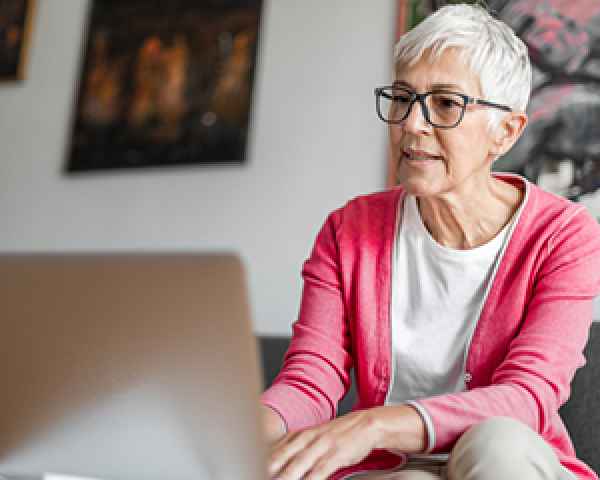 Senior citizen woking on a laptop in her living room and wearing a pink sweater and sitting on a chair 