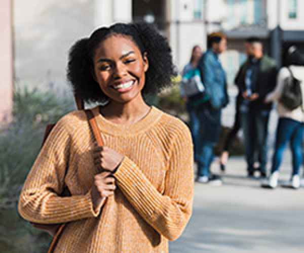 New college student stands outside on front of a building with students in the background. She is wearing a tan sweater and has a backpack over her shouldder. 