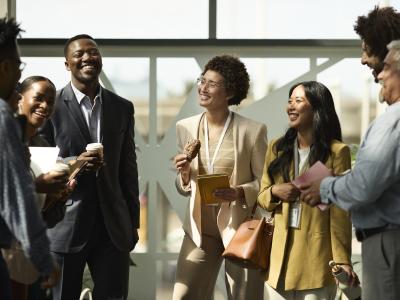 Group of business professionals standing in front of a window in a workplace environment. They are professionally dressed and talking and laughing with each other. 