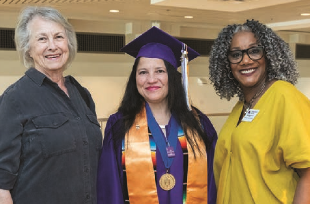 Commencement speaker stands in cap and gown next to two women and smile for the camera